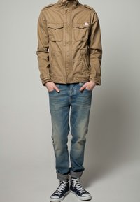 Brązowa kurtka na zamek z dwoma kieszeniami na piersi, zestawiona z jasnoniebieskimi dżinsami i granatowymi wysokimi trampkami z białymi sznurówkami i podwiniętym mankietem.