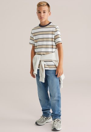Camiseta a rayas en colores beige, blanco y marrón; sudadera gris claro atada a la cintura; vaqueros de corte regular en azul; zapatillas grises con detalles en negro.