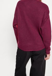 Burgunderfarbener Strickpullover mit lockerer Passform, runder Ausschnitt und kontrastierenden roten Einsätzen an den Seiten. Weicher Stoff, überschnittene Schultern.