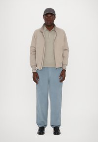 Beige Bomberjacke über einem hellgrauen Poloshirt, kombiniert mit lockeren hellblauen Hosen und schwarzen Schuhen. Eine graue Kappe vervollständigt das Outfit.