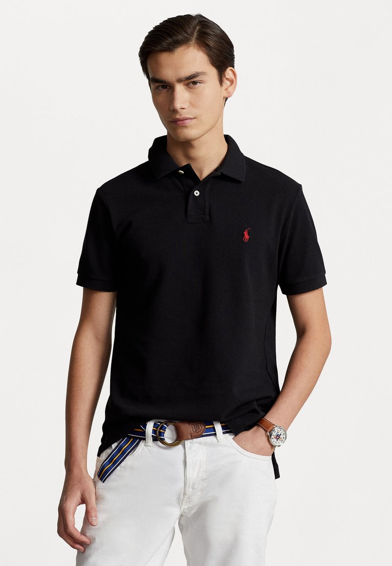 Svart polo-skjorte med korte ermer, knappet krage og rødt logo. Båret med hvite bukser og et brunt belte med stripete aksent.