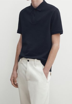 Polo-shirt navy à manches courtes, col classique et patte de boutonnage à deux boutons, associé à un pantalon couleur crème. Tissu lisse, coupe ajustée.