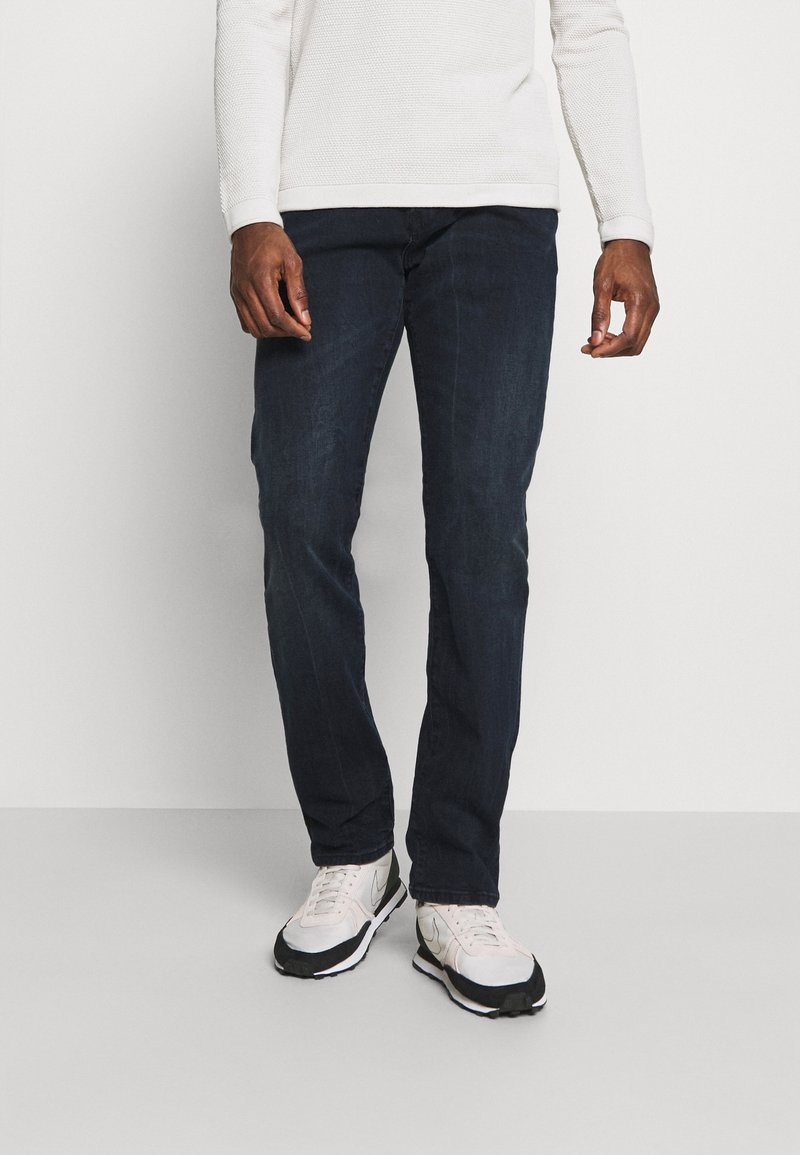 Homme portant un jean bleu foncé, une chemise blanche texturée à manches longues et des baskets blanches, debout devant un fond blanc uni.