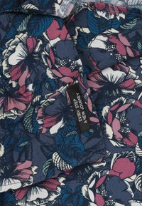 Bloemstof met een donkere marineblauwe achtergrond, voorzien van grote roze en witte bloemen en groene bladeren. Inclusief een zwart label met witte tekst.