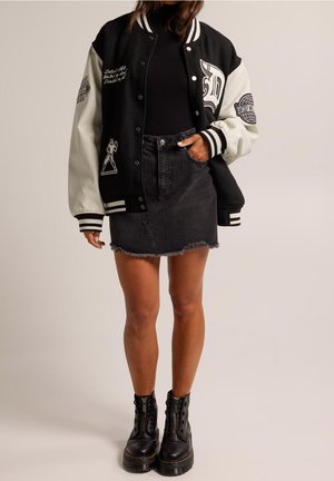 Chaqueta universitaria en blanco y negro con detalles bordados, combinada con una falda de mezclilla negra y botas negras con cordones. Tejido texturizado y mangas en contraste.