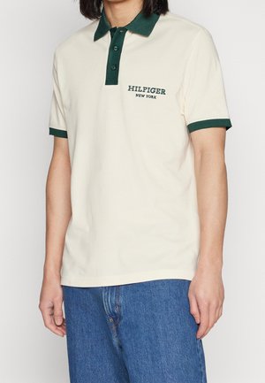 Osoba ubrana w kremową koszulkę polo z ciemnozielonym kołnierzykiem i brzegami rękawów, niebieskie dżinsy oraz tekst "HILFIGER NEW YORK" na klatce piersiowej.