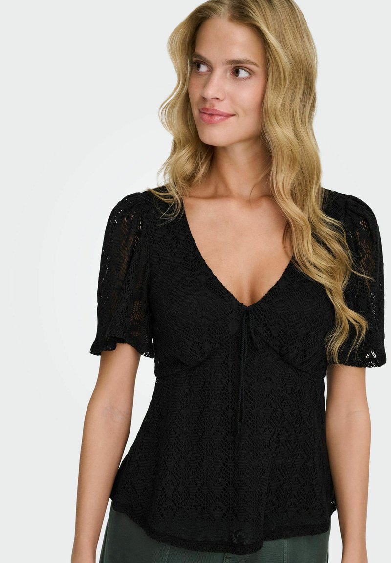 Femme aux longs cheveux blonds portant un haut court en dentelle noire à manches courtes avec un col en V et un détail subtil à nouer, regardant légèrement sur le côté.