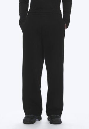 Person som har på seg løse svarte bukser, svart langermet topp og svarte treningssko, stående med hendene i lommene.