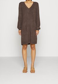 Robe marron en tricot avec un col en V et des manches longues. Texture ressemblant à un pull, longueur mi-cuisse, petites fentes latérales, portée avec des sandales à brides couleur sable.