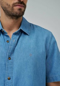 Camisa de hombre de manga corta con botones en denim azul claro. Cuenta con un cuello clásico, botones marrones y un pequeño logotipo bordado en el pecho.