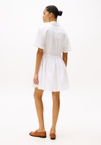 Rochie scurtă albă din in, cu guler și talie elastică, cu mâneci scurte și fustă evazată. Pantofii din piele maro completează look-ul.