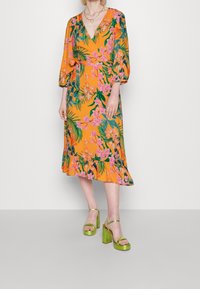 Kwiatowa sukienka z pomarańczowym tłem, ozdobiona różowymi kwiatami i zielonymi liśćmi, z dekoltem w kształcie V i bufkami; zestawiona z zielonymi platformami.