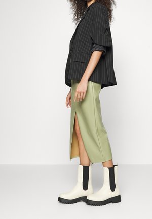 Mujer con blazer negro de rayas finas, falda midi verde claro con abertura frontal y botines blancos gruesos con laterales elásticos negros.