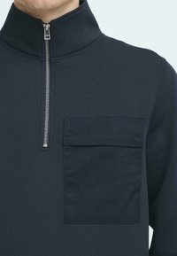 Navy blue pullover with a high collar, zippered front, and a chest pocket. Made from a smooth, textured fabric. Minimalist design.