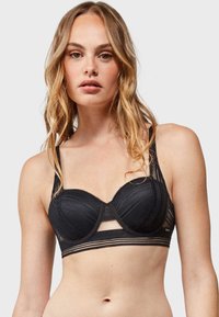 Sostén negro con copas moldeadas, acentos de malla transparente, bordes con forma de ondas y una banda con rayas horizontales. Tirantes ajustables y cierre de gancho.
