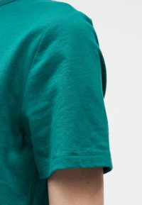 Κοντινό πλάνο του άνω βραχίονα ενός ατόμου που φορεί μια τιρκουάζ κοντομάνικη μπλούζα πάνω σε ανοιχτό, μονόχρωμο φόντο.