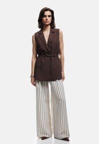 Blazer senza maniche marrone con vita cintura, abbinato a pantaloni crema a gamba larga con strisce verticali nere e calzature marroni a punta.