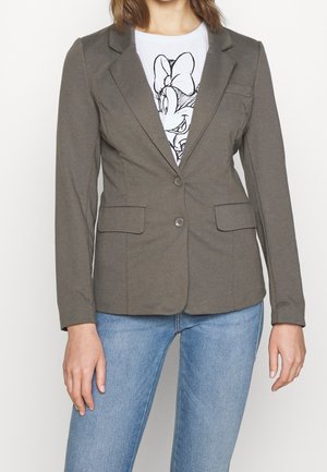 Femme portant un blazer gris cintré sur un t-shirt blanc à motif Minnie Mouse et un jean bleu clair, debout devant un fond uni.