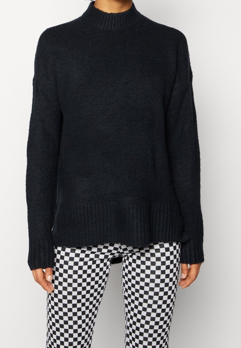 Personne portant un pull en maille noire avec ourlet et manches côtelées, associée à un pantalon à carreaux noir et blanc, debout devant un fond uni.