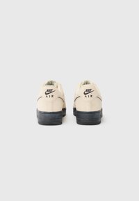 Nike Air sneakers har en lys beige overdel med en tekstureret finish, sort sål og broderet branding på hælen.