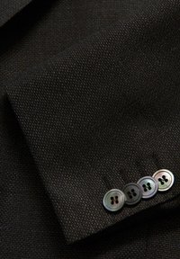 Manica in tessuto nero con una trama testurizzata, caratterizzata da quattro bottoni iridescenti al polsino, messi in risalto da cuciture a contrasto.