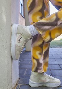 Korkease valkoiset lenkkarit vaaleanharmailla yksityiskohdilla ja mustalla swooshilla, paritettu kuviollisten keltaisten ja ruskeiden housujen kanssa, nojaamassa seinää vasten.