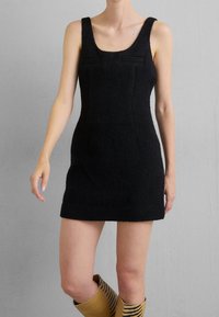 Schwarzes gestricktes Minikleid mit breiten Trägern, rundem Ausschnitt und vorderen Taschen. Figurbetontes Design und strukturierter Stoff, getragen mit gemusterten kniehohen Stiefeln.