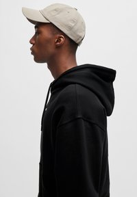 Zwarte hoodie met een grote capuchon, gemaakt van katoen, met een voorzak. Beige pet op het hoofd, met een gebogen rand en stikdetails.