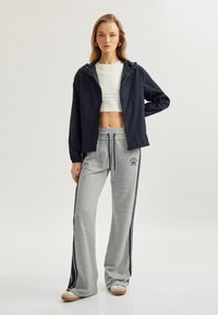 Marinfärgad lätt huva jacka över en vit kort topp, kombinerad med gråa sweatpants med sidostripes och logotypdetalj, på en modell.