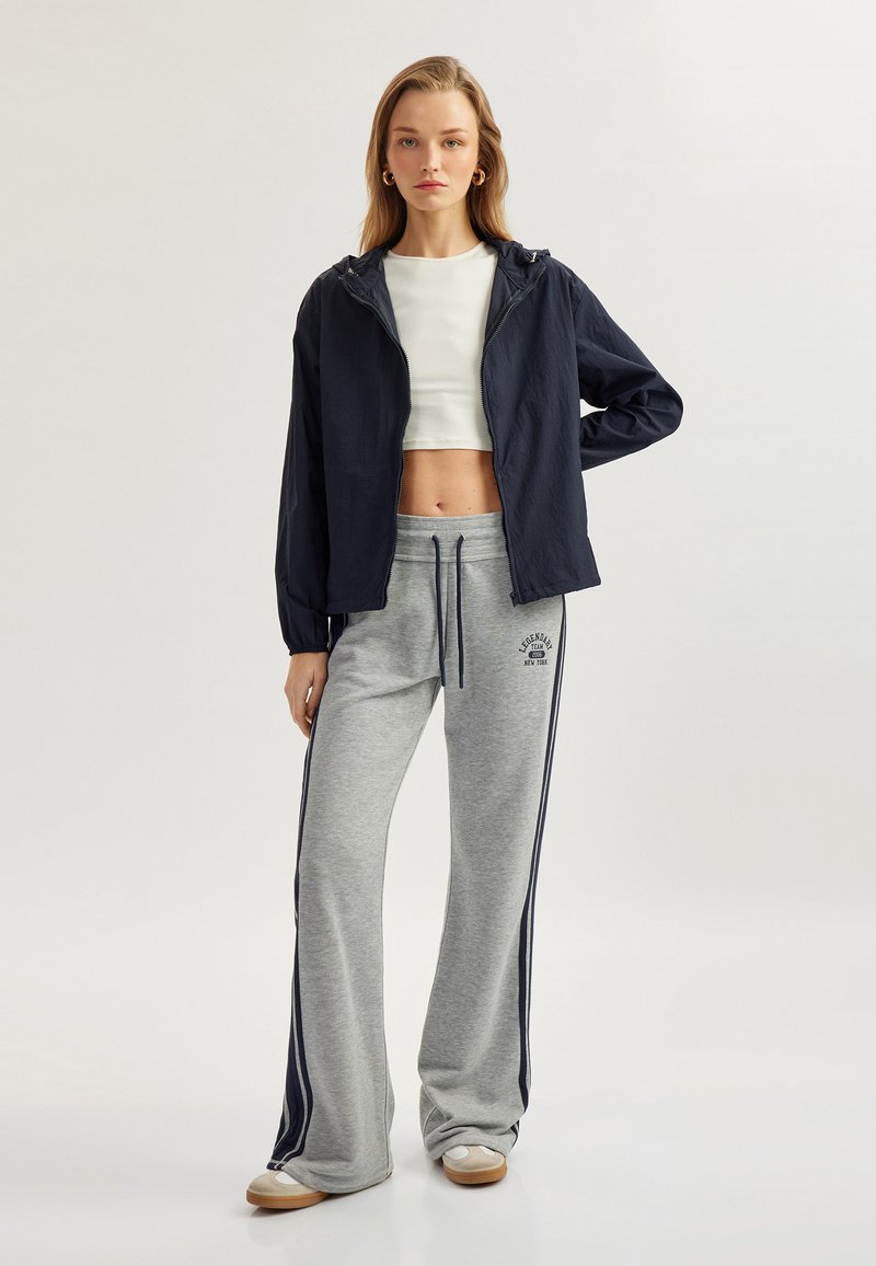 Marinfärgad lätt huva jacka över en vit kort topp, kombinerad med gråa sweatpants med sidostripes och logotypdetalj, på en modell.