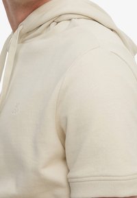 Felpa con cappuccio beige dal texture morbido. Presenta una tasca a marsupio e un logo ricamato sul petto. Maniche corte e vestibilità comoda.