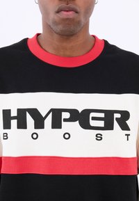 Čierno-biela bavlnená tričko s červenými akcentmi. Obsahuje výrazný text "HYPER BOOST" cez prsia. Kolo výstrih a jemný povrch.