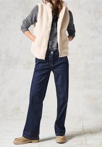 Beige Fleece-Weste über einem grauen Strickpullover, kombiniert mit dunkelblauen weiten Jeans und hellen beigen Slippern. Weiche Texturen und lässiges Design.