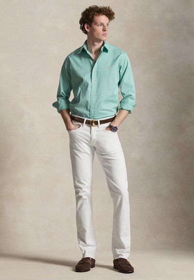 Νέος άνδρας στέκεται με τα χέρια στις τσέπες, φοράει πουκάμισο μέντας με κουμπιά, λευκό παντελόνι, καφέ ζώνη, ρολόι και καφέ παπούτσια, μπροστά σε απλό φόντο.