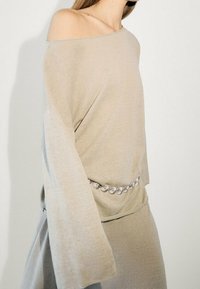 Beige, schulterfreies Oberteil mit langen, weiten Ärmeln und einer sanften Textur, versehen mit einem silbernen, runden Gürtelakzent in der Taille.