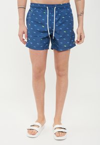 Pantaloni da bagno blu con un motivo ripetuto di piccole forme verdi e blu, vita regolabile con cordino e tasche laterali. Indossati con sandali bianchi.