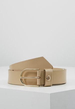 Beiger Ledergürtel mit einer goldenen, runden Schnalle und einem kleinen goldenen Emblem an der Gürtelschlaufe, auf einer weißen Oberfläche präsentiert.