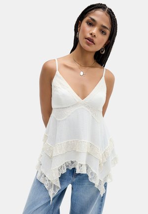 Donna con capelli intrecciati che indossa un top bianco in pizzo a balze e jeans blu, accessoriata con orecchini a cerchio dorati e collane a più strati.