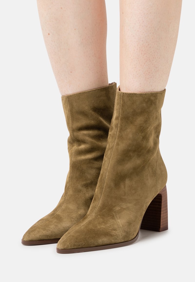 Bottes mi-mollet en daim marron avec bouts pointus et talons blocs, portées sur des jambes nues, présentées sur un fond clair uni.