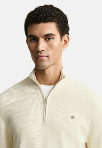 Cremfarbener Reißverschluss-Pullover mit strukturiertem Strickdesign, ausgestattet mit einem Viertel-Reißverschluss und einem kleinen Logo auf der linken Brust.