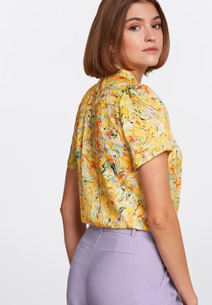Blusa a maniche corte gialla con motivi a vortice. Abbinata a pantaloni su misura viola chiaro, caratterizzati da una superficie liscia e linee pulite.