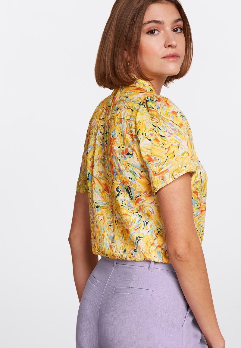 Koszulka na krótki rękaw w żółtym kolorze z wzorami w formie wirów kolorów. Połączona z jasnofioletowymi spodniami o dopasowanym kroju, które mają gładką teksturę i czyste linie.