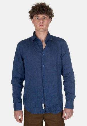 Uomo con capelli corti e ricci indossa una camicia blu a maniche lunghe abbottonata e pantaloni marroni, in piedi rivolto in avanti contro uno sfondo semplice.