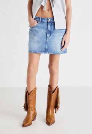 Hellblaue Denim-Minirock mit vorderen Taschen, akzentuiert durch einen Metallknopf und dezente Nähte. Kombiniert mit braunen Cowboy-Stiefeln mit Fransen.