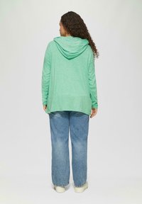 Grüne Kapuzenjacke aus leichtem, strukturiertem Stoff, mit Kapuze und lockerer Passform. Kombiniert mit lockeren blauen Jeans.