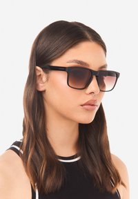 Mustat suorakulmaiset aurinkolasit mattapintaisella viimeistelyllä ja liukuvärjätyillä ruskeilla linsseillä. Ominaisuuksiin kuuluvat paksut kehykset ja hillitty brändäys sangan kohdalla.