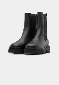 Par svart lær ankelstøvler med tykke knotter såler og elastiske sidepaneler på en ensfarget lys bakgrunn.