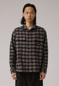 Camicia di flanella con un motivo a quadri marrone e blu navy, bottone frontale, due tasche sul petto e un logo a patch sulla tasca sinistra.