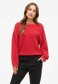 Maglione rosso con una texture morbida, vestibilità rilassata e maniche lunghe. Presenta un collo a girocilo accentuato da piccoli dettagli decorativi. Abbinato a pantaloni neri.