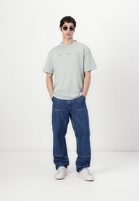 Mintgroen oversized T-shirt met korte mouwen, gecombineerd met losse blauwe denim broek. Witte sneakers en ronde zonnebril maken de look compleet.